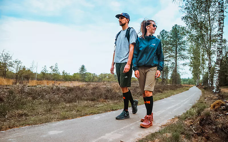 Twee mensen wandelen over een verhard pad door een natuurgebied met bomen en gras. Ze dragen outdoorkleding, rugzakken en kniekousen en genieten van een zonnige dag in de buitenlucht.